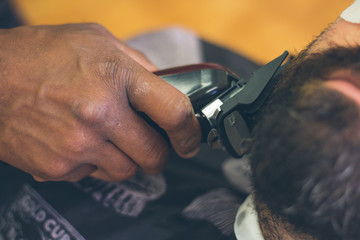 Closeup view of barber's hand using trimmer to cut the beard of his client at barbershop.