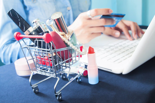 Cosmetics and woman's essentials in shopping cart with woman using laptop and credit card for online shopping