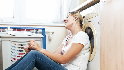 Fototapeta na wymiar Portrait of young smiling woman sitting on floor at laundry and listening music with earphones