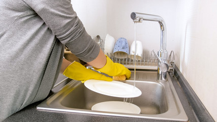 Young housewife in yellow rubber gloves washing plates in kitchen sink