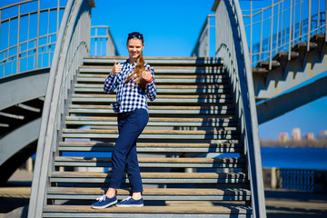 Young stylish girl with thumbs up stands on street stairs in sunny day. Street style