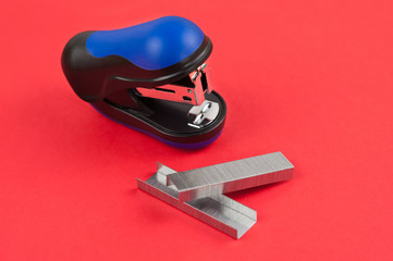Two stationery new metal clips and black and blue stapler on red background