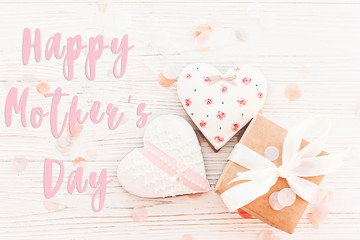 happy mother's day text on cookie hearts and gift box on white rustic wooden background with confetti in soft light. flat lay. stylish mothers day greeting card, top view. cookie for mom