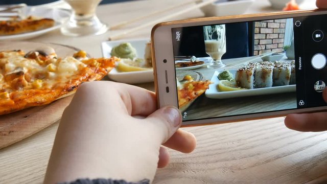 Hands of a teenager make a photo of food on a smartphone. A set of sushi rolls from Japanese cuisine and pizza on the background of porcelain teapot for soy sauce and saucer in a stylish cafe