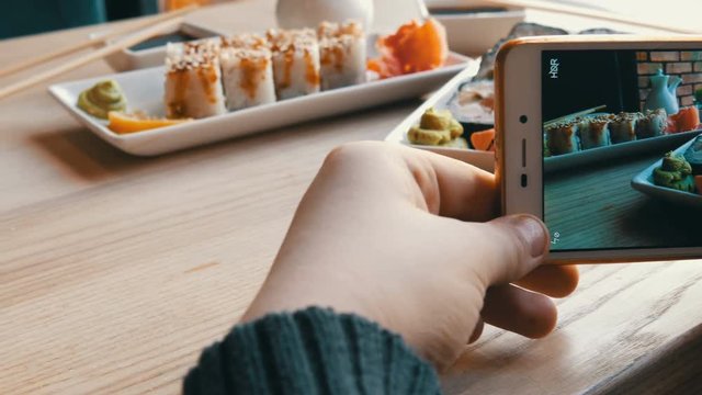 Hands of a teenager make a photo of food on a smartphone. A set of sushi rolls from Japanese cuisine on the background of porcelain teapot for soy sauce and saucer in a stylish cafe