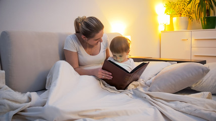 Happy young woman reading story to her toddler son in bed at night