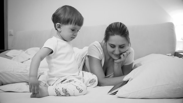 Black and white image of cute toddler boy with young mother watching cartoons on tablet in bed