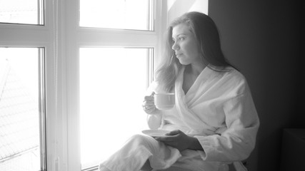 Black and white image of young woman in bathrobe sitting on windowsill and drinking tea