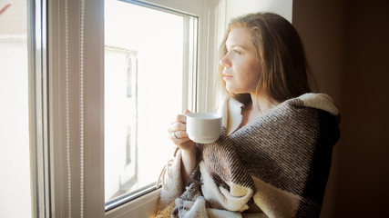 Closeup toned portrait of smiling young woman covering in plaid with cup of tea standing at window