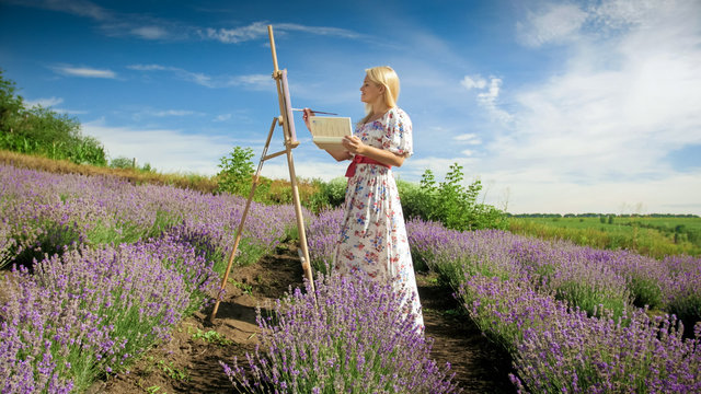 Beautiful young woman in long dress making painting of lavender field in Provence