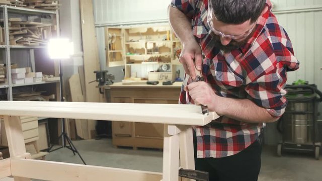 man carpenter in a shirt with a beard in the workshop is working on a wooden bench