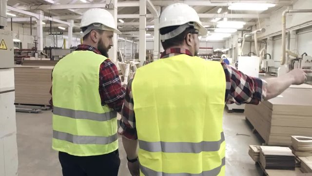 two workers talking in green waistcoats and helmets at a furniture factory among machines
