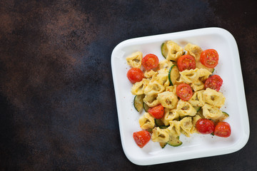 Tortellini with roasted zucchini and cherry tomatoes served on a white plate, above view on a dark brown metal background with copyspace