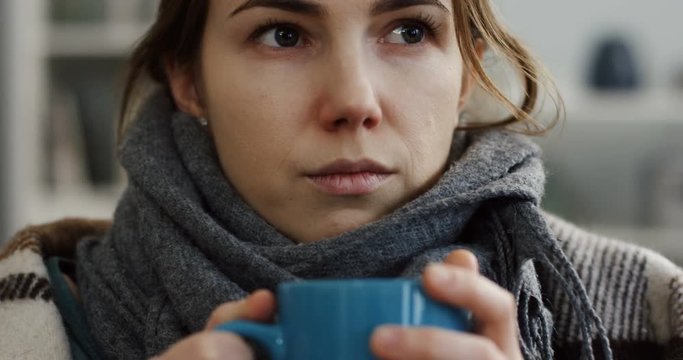 Young caucasian unwell woman in the warm scarf sitting coverd with a plaid and drinking hot tea as having a flu. At home. Indoors
