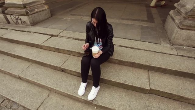 Beautiful girl with dark hair, black jacket, white sneakers, with coffee, browsing news and social networks in her smartphone.