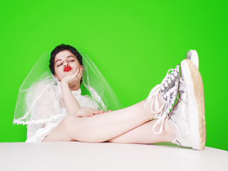 A beautiful bride is smiling, sitting with her feet on the table and feeling happy for the sake of anticipation of a merry and main party in life.Green background