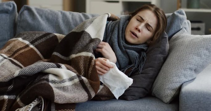 Unhealthy young woman in a scarf and a plaid sneezing in a napkin while lying on the sofa under the plaid in the living room, having a bad cold. Inside