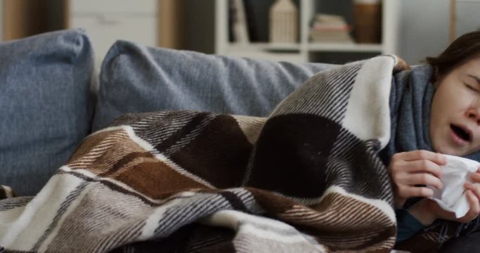 Unwell young woman in a scarf and covered with a plaid sneezing in a napkin while lying on the gray couch in the cozy living room, having a flu. Indoors