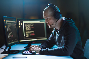 cybercrime, hacking and technology concept - male hacker with headset and coding on computer screen...