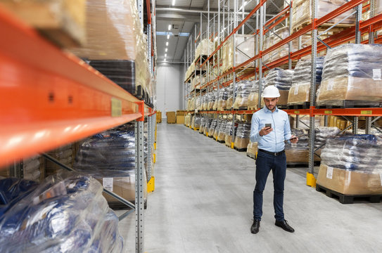 wholesale, logistic business, technology and people concept - businessman in helmet with smartphone and clipboard at warehouse