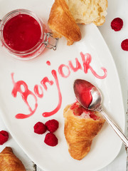 Close view of fresh croissants served with ripe raspberries and raspberry jam on white plate with lettering Bonjour over wooden surface. © PINKASEVICH