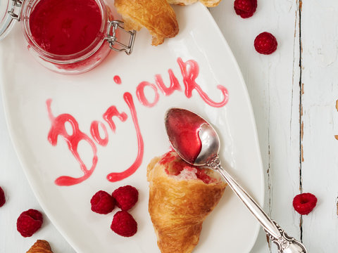 Close view of fresh croissants served with ripe raspberries and raspberry jam on white plate with lettering Bonjour over wooden surface.