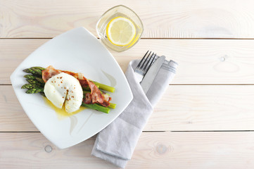 On a plate of asparagus with slices of fried bacon. On top of the asparagus is an egg poached. Next napkin, cutlery and a glass of water. Light wooden background. View from above. Close-up.