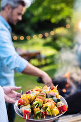 In summer. A couple prepares a bbq to welcome friends in the garden. Close-up on a plate of grill skewers - obrazy, fototapety, plakaty