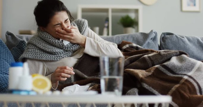 Close up of the blurred lemon, medicines, pills, glass of water and drops on the table. Ill woman lying under the plaid on the couch and caughing on the background. Inside
