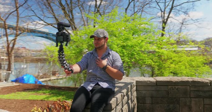 A young millennial vlogger sits on a stone wall dangling his feet while recording a daily video for his social media channel.  	