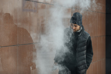 Vape teenager. Portrait of a handsome young white guy in black waistcoat and modern cap vaping an electronic cigarette opposite the futuristic modern background. Lifestyle.