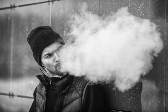 Vape teenager. Portrait of a handsome young white guy in black waistcoat and modern cap vaping an electronic cigarette opposite the futuristic urban background. Black and white.