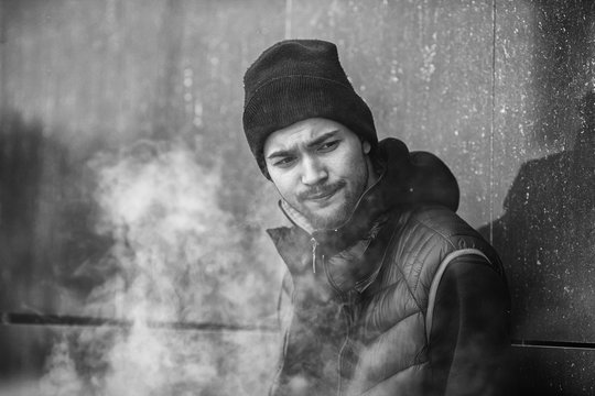 Vape teenager. Portrait of a handsome young white guy in black waistcoat and modern cap vaping an electronic cigarette opposite the futuristic urban background. Black and white.