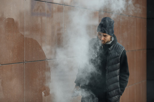 Vape teenager. Portrait of a handsome young white guy in black waistcoat and modern cap vaping an electronic cigarette opposite the futuristic modern background. Lifestyle.