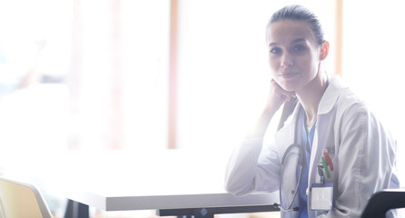 Naklejka na ściany i meble Beautiful young smiling female doctor sitting at the desk. Woman doctor