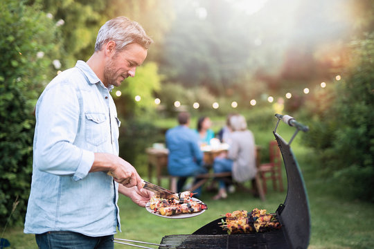 in the summer,  a handsome man in his forties prepares a barbecue for his friends gathered around a table in the garden