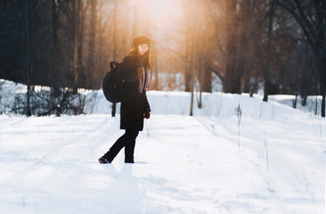 A young original girl in a hat with a visor and a black backpack walks through the winter clearing in the rays of the evening sun and looks into the frame at the viewer.