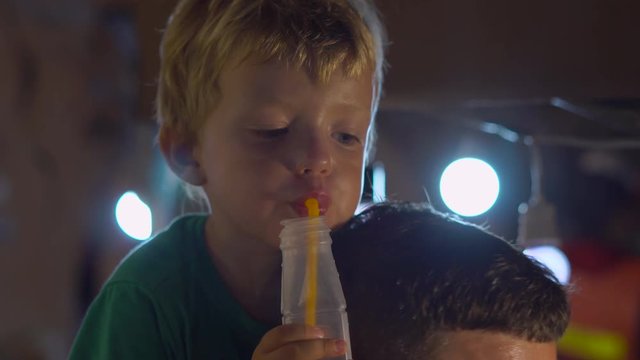 Little boy dring fresh fruit juice sitting on his father's neck on a night market in Thailand. Thai food concept. Asian food concept