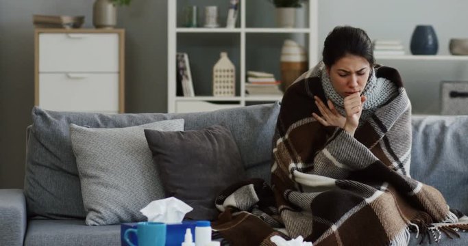 Caucasian young unhealthy woman sitting on the gray couch in the living room covered with a plaid having a severe caugh as she having a flu. Indoors