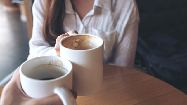 Close up image of a man and a woman clinking white coffee mugs in cafe