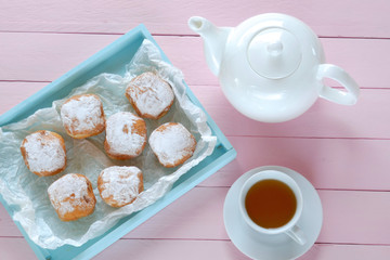 Morning tea flat lay. sweet pastries. donuts with powdered sugar in a blue tray,  white kettle and a cup of tea on a pink wooden plank background. flat lay in pastel colors. top view,