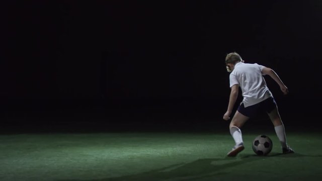 Junior soccer player exercising with ball on artificial turf in dark indoor arena with low key lightning