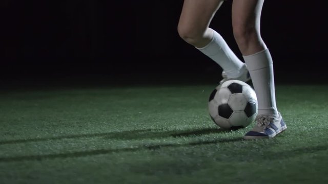 Slow motion shot of legs of junior soccer player practicing dribbling technique with ball on artificial turf in dark indoor arena