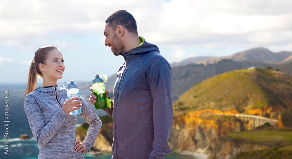 Wall mural fitness, sport and people concept - smiling couple with bottles of water over bixby creek bridge on big sur coast of california background