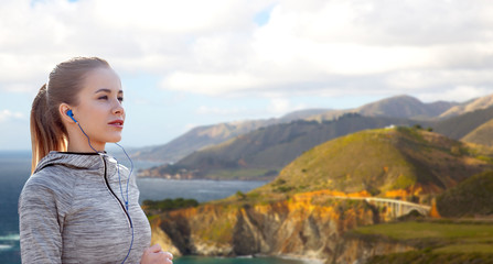 fitness, sport and technology concept - happy woman running and listening to music in earphones over bixby creek bridge on big sur coast of california background