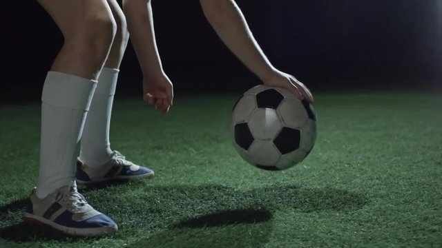 Low section of junior soccer player dribbling a ball with hand and then kicking it on field with artificial turf in dark arena