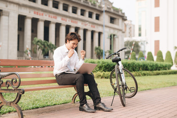 Young handsome businessman with laptop and bicycle sitting on bench in park