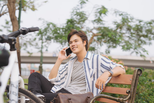 Young handsome businessman talking by mobile phone while sitting on bench in park after riding bicycle