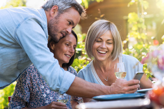 In summer. Group of friends gathered around a table in the garden to share a meal. a gray-haired man makes them laugh by showing them pictures on his phone.