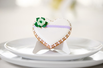 Heart shaped glazed cookie - decorated with green glaze flower and tiny pattern stands on a white plates as a decoration for festive wedding table. A cookie stands on the white restaurant background.